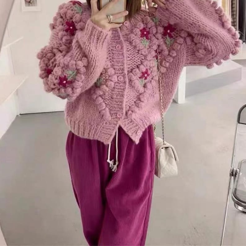 Puede incluir: Un c&aacute;rdigan de punto rosa con un dise&ntilde;o floral y pompones. El c&aacute;rdigan tiene cierre de botones y mangas largas.