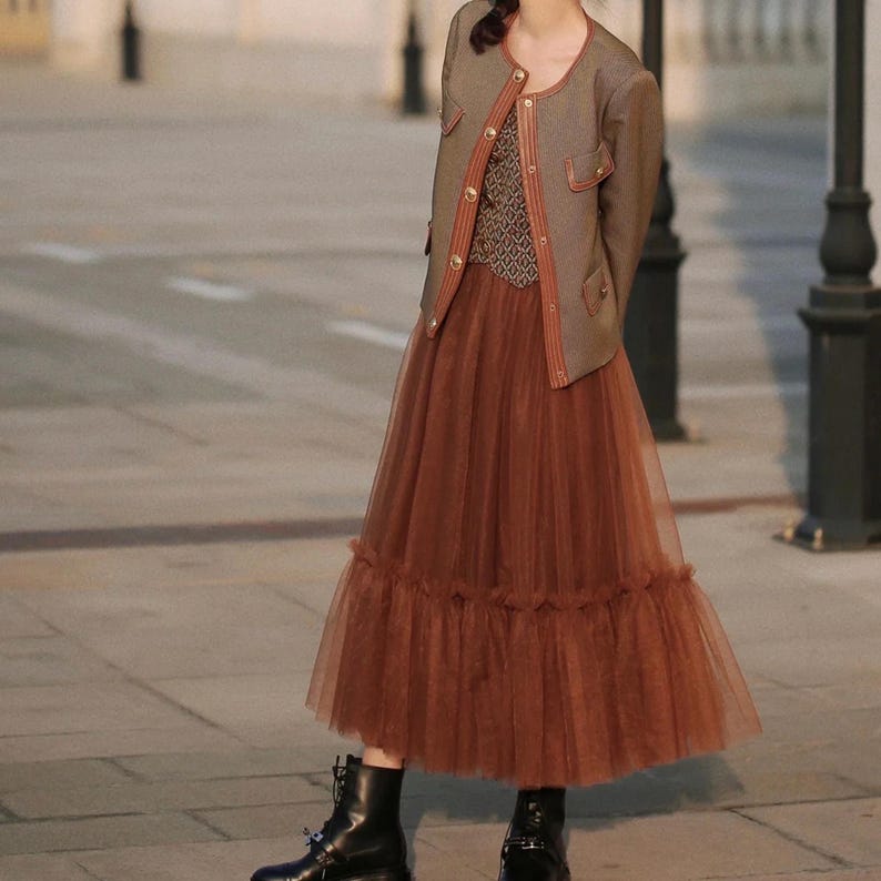 May include: A woman wearing a brown jacket with gold buttons over a patterned top and a long brown tulle skirt with a ruffled hem. She is wearing black boots.