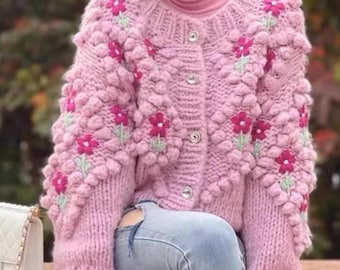 May include: A pink knitted cardigan with a button closure and embroidered flowers. The cardigan has a textured, puffy design.