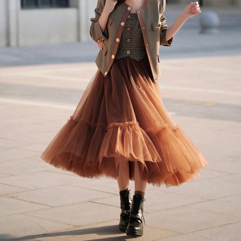 May include: A brown tulle skirt with multiple layers and a ruffled hem. The skirt is paired with a brown jacket and a patterned vest.