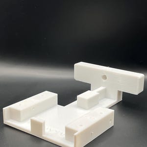 Peut inclure: Base en plastique blanc imprimée en 3D avec une pièce en forme de T sur le dessus. La base comporte quatre fentes rectangulaires et une petite pièce rectangulaire au centre. La pièce en forme de T a un trou au centre.