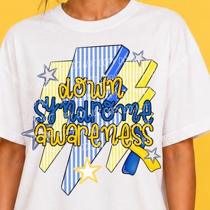 May include: A white t-shirt featuring a graphic design with blue and yellow lightning bolt shapes, stars, and the text "down syndrome awareness" in a yellow font with a blue outline. The design includes blue and white vertical stripes.