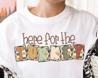 May include: White crew neck t-shirt featuring the text "here for the TURKEY" in a decorative font. The word "TURKEY" is in colourful, patterned blocks. The shirt is part of a Thanksgiving or autumn-themed collection.