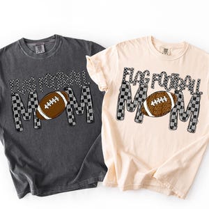 May include: Two t-shirts, one charcoal and one cream, feature the text "FLAG FOOTBALL MOM" in a checkered pattern. A brown football graphic is centered within the word "MOM". The shirts are short-sleeved and have a crew neck.