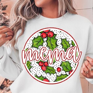 May include: White sweatshirt with a round graphic featuring the word "mema" in red script. The graphic includes green holly leaves, red berries, and a dotted background, all within a red circle. A festive holiday design.
