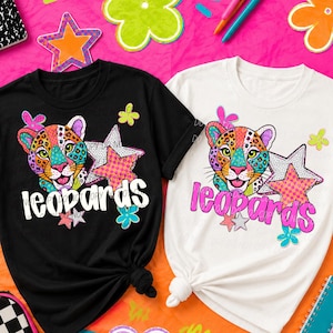 May include: A black t-shirt and a white t-shirt, each featuring a colourful graphic of a leopard face with a patterned star, small flowers, and the word leopards printed in a bold, stylised font across the centre of the chest area.