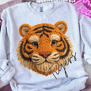 May include: A light gray sweatshirt featuring a detailed, textured tiger face in shades of orange, brown, and black. The word "Tigers" is written in a black script font below the tiger's face. The sweatshirt has a crew neck and long sleeves.