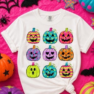 May include: A white short-sleeve t-shirt featuring a graphic print of nine colorful jack-o'-lantern pumpkins arranged in a grid. Each pumpkin displays a unique facial expression and pattern, including stripes, stars, and polka dots in vibrant neon colors.