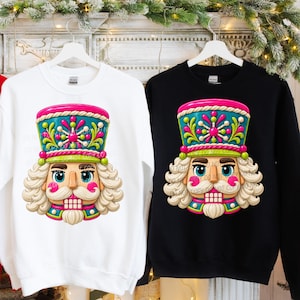 May include: Two crewneck sweatshirts, one white and one black, each featuring a colorful graphic of a nutcracker face. The nutcracker has a teal hat with pink and white accents, a white mustache, a white beard, and a pink-cheeked expression.