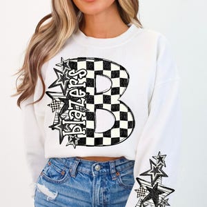 Puede incluir: Sudadera blanca con la palabra "Blazers" en un diseño en forma de estrella y una gran "B" a cuadros en la parte delantera. La manga izquierda presenta un grupo de estrellas y gráficos de banderas a cuadros. La sudadera se combina con vaqueros azules.