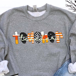 May include: A heather gray crewneck sweatshirt featuring the word teacher in a decorative font. The letters alternate between orange and white candy corn stripes, black with white polka dots, and black with a subtle spider web pattern.