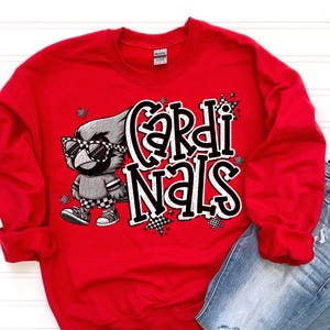 Puede incluir: Una sudadera roja con la palabra "Cardinals" en una fuente estilizada, acompañada de un cardenal de dibujos animados con gafas de sol. El cardenal viste un atuendo a cuadros grises y negros. La sudadera se combina con vaqueros azules.