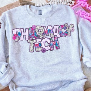 Może przedstawiać: Szara bluza z napisem "Pharmacy Tech" w różowych literach z kwiatowym i kratkowym wzorem.