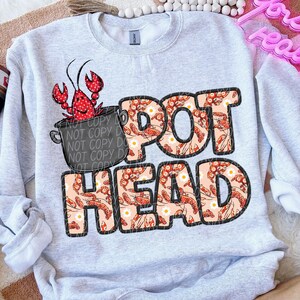 May include: A gray sweatshirt with a design of a pot of crawfish with the text "POT HEAD" in red and white letters. The letters are filled with a pattern of crawfish and white flowers.