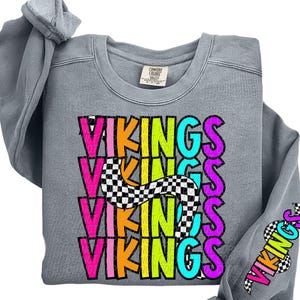 May include: A gray sweatshirt with the word "VIKINGS" in a stacked, colorful design. The text is in neon pink, yellow, orange, and teal, with a checkered pattern accent. The sleeve also has the word "VIKINGS" in a similar style.