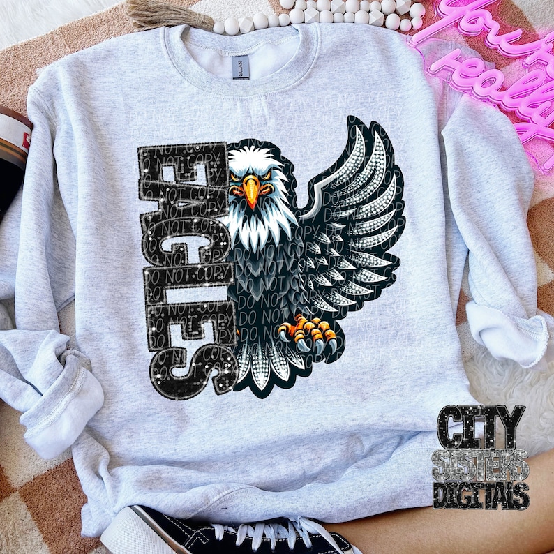 Peut inclure: Un sweat-shirt gris avec un motif d'aigle en strass noir et blanc avec le mot "Eagles" en strass. L'aigle a les ailes d&eacute;ploy&eacute;es et fait face &agrave; droite.