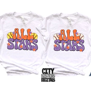 Pu&ograve; includere: Due magliette bianche con il testo "All Stars" in un font retr&ograve; viola e arancione. Il testo &egrave; circondato da palline da baseball e stelle.
