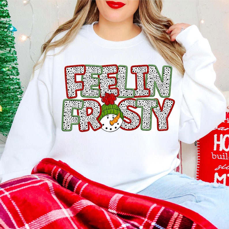 May include: White sweatshirt with the words "FEELIN FROSTY" in a polka dot design with a snowman graphic. The text is outlined in red and green. The snowman is wearing a green hat with a red pom-pom and has an orange carrot nose.