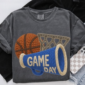 May include: Dark grey long-sleeve shirt with a basketball, net, and megaphone design. The blue megaphone has "GAME DAY" in white letters. The basketball is brown and the net is tan.