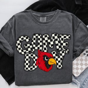 May include: Dark gray sweatshirt with the words "GAME DAY" in a checkered pattern, and a red cardinal graphic. The sweatshirt has long sleeves and a crew neck. A phone with a checkered case is visible.