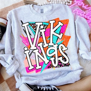 May include: A light gray sweatshirt with the word "VIKINGS" in large white letters. The letters are outlined in black and have a colorful background with pink, orange, and teal accents. The sweatshirt has long sleeves.