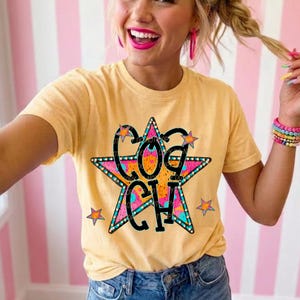 May include: A light yellow short-sleeve t-shirt featuring a large, multi-colored star graphic with a teal outline and pink accents. The word COACH is printed in bold black letters across the center of the star, surrounded by smaller stars.