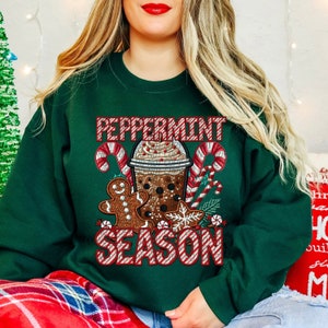 Puede incluir: Sudadera verde oscuro con un gr&aacute;fico festivo. El gr&aacute;fico presenta una bebida navide&ntilde;a, hombre de jengibre, bastones de caramelo y las palabras "Peppermint Season" en rojo y blanco. La sudadera es una prenda de vestir de temporada y acogedora.