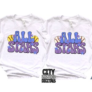 Pu&ograve; includere: Due magliette bianche con le parole "All Stars" in lettere viola e blu con una grafica di softball. Le magliette sono su uno sfondo bianco.