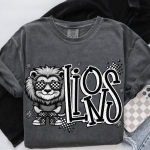 May include: Dark gray sweatshirt featuring a cartoon lion wearing checkered sunglasses and matching shoes. The word "LIONS" is printed in a bold, stylized font with checkered accents. A phone case with a checkered pattern is visible.