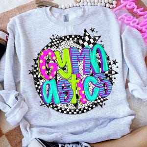 Peut inclure: Un sweat-shirt gris avec un motif graphique color&eacute; qui dit "Gymnastics" dans un cercle avec des &eacute;toiles et des motifs &agrave; carreaux.