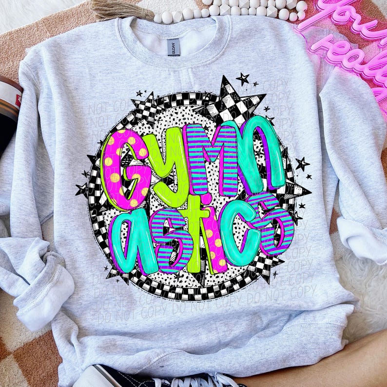 Peut inclure: Un sweat-shirt gris avec un motif graphique color&eacute; qui dit "Gymnastics" dans un cercle avec des &eacute;toiles et des motifs &agrave; carreaux.