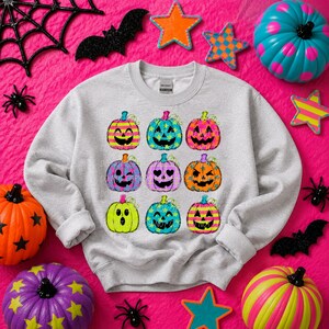 May include: A light gray crewneck sweatshirt featuring a graphic print of nine colorful jack-o'-lanterns in three rows. The pumpkins display various patterns including stripes, stars, and polka dots in vibrant neon colors like pink, blue, and yellow.