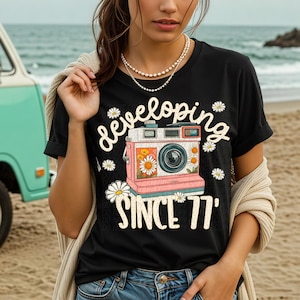 May include: Black t-shirt with a retro camera graphic and the text "developing since '77". The camera is pink, white, and orange, decorated with daisies. The shirt is a classic crew neck.
