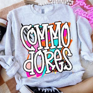 May include: A light gray sweatshirt with the word "Commodores" in large white, graffiti-style lettering. The lettering is layered over a colorful abstract design with orange, pink, and teal accents. The sweatshirt has a crew neck and long sleeves.