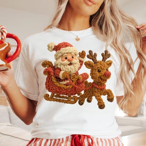 May include: White t-shirt featuring a festive design of Santa Claus in a sleigh pulled by a reindeer. The Santa and reindeer are in shades of brown, red, and beige, perfect for the holiday season.