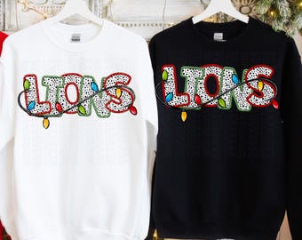 May include: Two sweatshirts, one white and one black, each displaying the word "LIONS" in a festive design. The letters are filled with a dalmatian-like pattern and outlined in green, with a string of colorful Christmas lights wrapped around them.