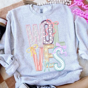 May include: A light gray sweatshirt with the word "WOLVES" in colorful, patterned letters. Each letter has a unique design, including floral, checkered, and polka dot patterns, and is adorned with decorative bows in various colors. The sweatshirt has a crew neck and long sleeves.