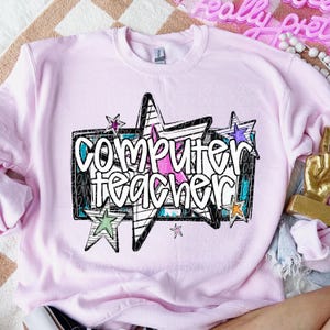 Op de afbeelding: Een lichtroze sweatshirt met de tekst "Computer Teacher" in het wit, omlijnd met zwart. De afbeelding bevat sterren in verschillende kleuren en een zwart-witte achtergrond.