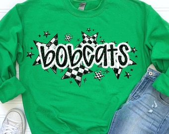Pode incluir: Uma camisola verde com a palavra "Bobcats" em branco, rodeada por estrelas xadrez em preto e branco e pequenas estrelas pretas. A camisola é mostrada com jeans azuis e tênis brancos.
