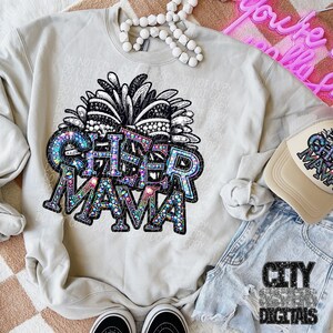 May include: A gray sweatshirt with a rhinestone design that says "Cheer Mama" with a black and white pom-pom on top. The sweatshirt is laying on a light brown surface with a pair of black sneakers, a pair of blue denim shorts, and a beige baseball cap with the same rhinestone design.