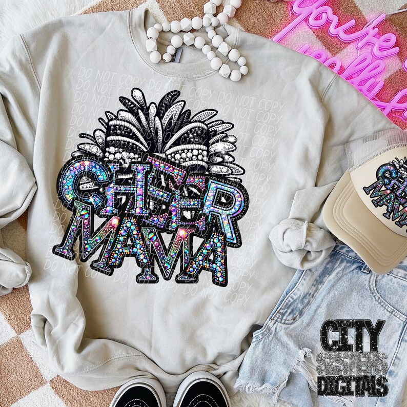 May include: A gray sweatshirt with a rhinestone design that says "Cheer Mama" with a black and white pom-pom on top. The sweatshirt is laying on a light brown surface with a pair of black sneakers, a pair of blue denim shorts, and a beige baseball cap with the same rhinestone design.