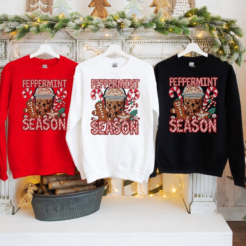 Puede incluir: Tres sudaderas en rojo, blanco y negro, cada una con un dise&ntilde;o festivo de "Peppermint Season". El gr&aacute;fico incluye una bebida de caf&eacute;, un hombre de jengibre, bastones de caramelo y acebo.