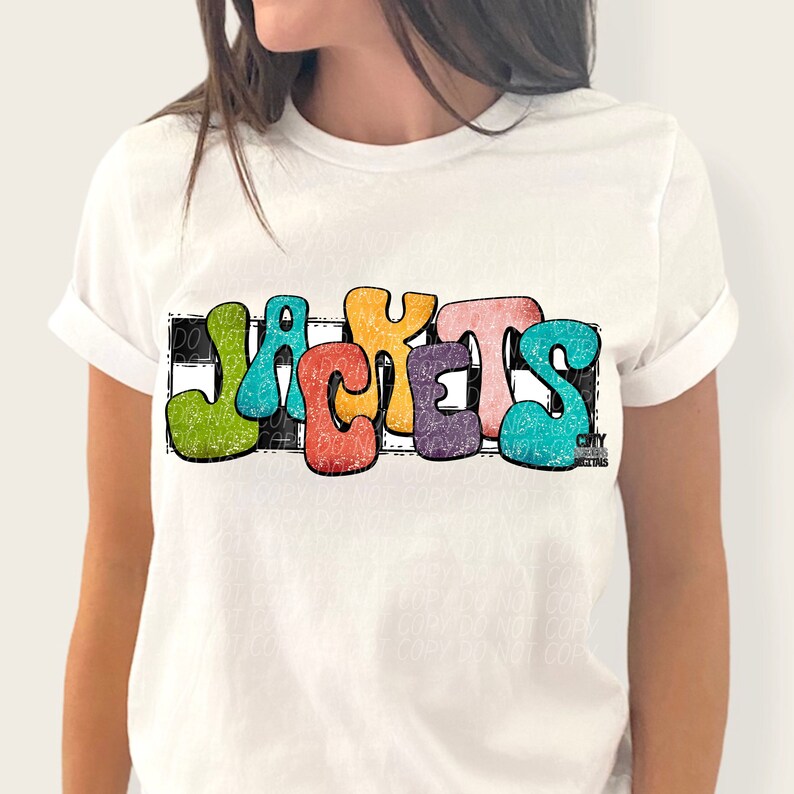 May include: A white t-shirt with a colorful graphic of the word "JACKETS" in a retro style. The letters are outlined in black and have a distressed look. The graphic is set against a black and white checkered background.