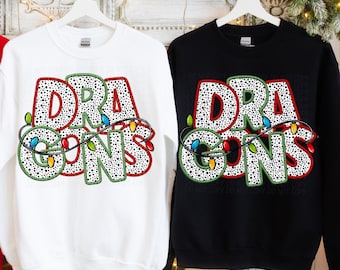 Puede incluir: Dos sudaderas, una blanca y otra negra, que muestran la palabra "DRAGONS" en un diseño festivo. Las letras están moteadas con puntos negros, delineadas en rojo y adornadas con una guirnalda de luces navideñas de colores.