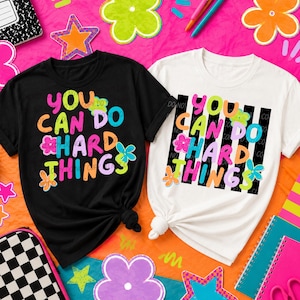 May include: A black t-shirt and a white t-shirt, both featuring the colorful text "YOU CAN DO HARD THINGS" in a playful font, accented with small flower graphics. The white shirt also includes a black and white vertical striped background design.