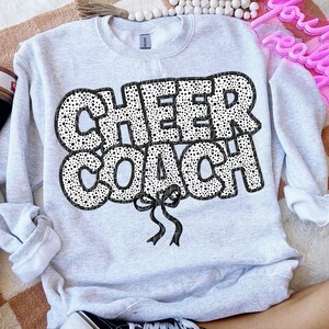 Puede incluir: Una sudadera gris con un dise&ntilde;o de lunares negros y blancos que dice "CHEER COACH" con un lazo negro.