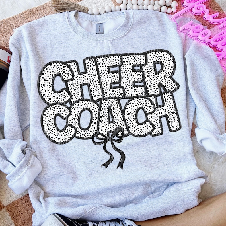 Puede incluir: Una sudadera gris con un dise&ntilde;o de lunares negros y blancos que dice "CHEER COACH" con un lazo negro.
