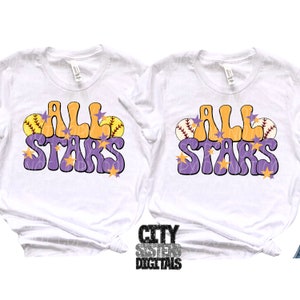 Pu&ograve; includere: Due magliette bianche con il testo "All Stars" in lettere viola e arancioni con grafica da softball. Il testo &egrave; in uno stile di font retr&ograve;.
