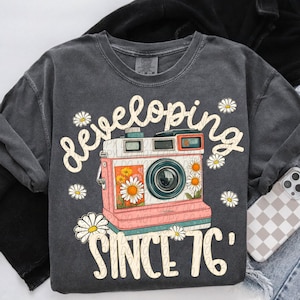 May include: Dark gray sweatshirt with a graphic of a vintage camera with the words "developing since '76". The camera is pink and white with a floral design and daisy accents. The sweatshirt is a comfortable, casual style.