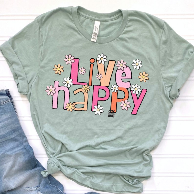 May include: Sage green t-shirt with the text "Live Happy" in pink, orange, and yellow letters. The letters are surrounded by white daisies with pink and yellow centers.